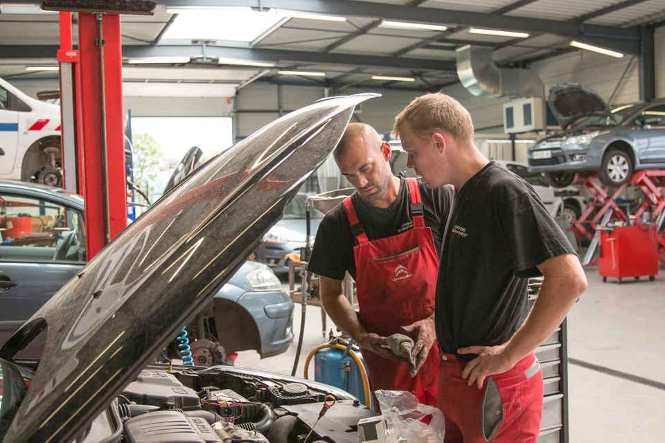 Deux techniciens en combinaison de travail bleue installent un pare-brise automobile dans un atelier de réparation vitré